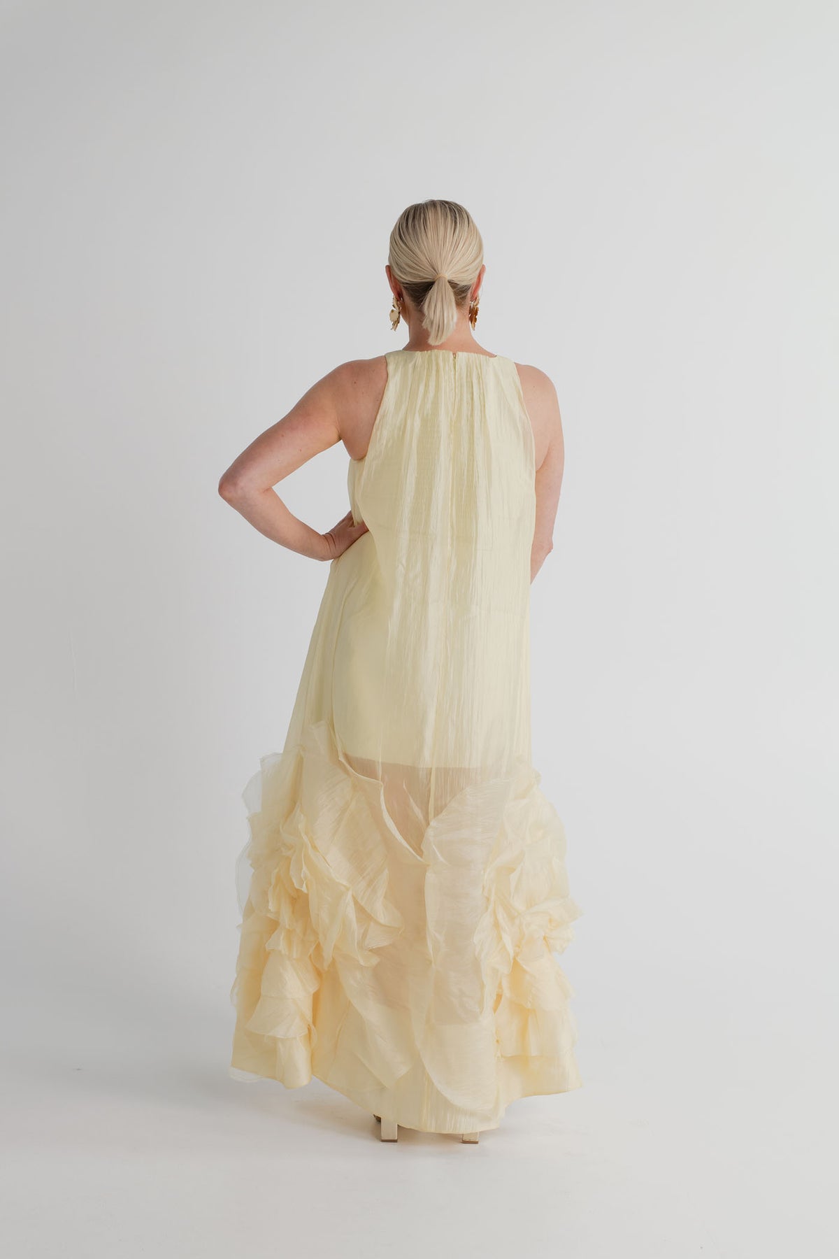 Back view of the Aje Ecliptica pastel yellow sleeveless gown showing its fluid shift silhouette and sculptural rosette hem.