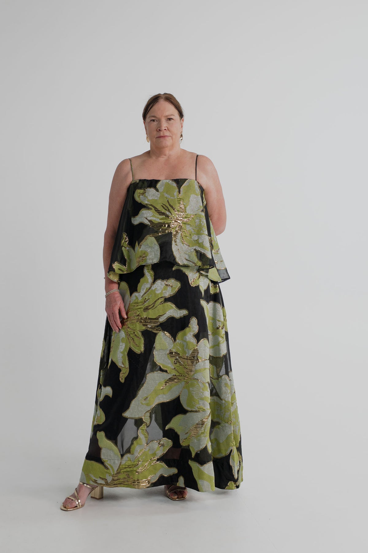Elliatt Elysia Tiered Gown in black with oversized green, silver and gold metallic floral detailing, thin straps and a layered tiered silhouette, shown full length from the front.

