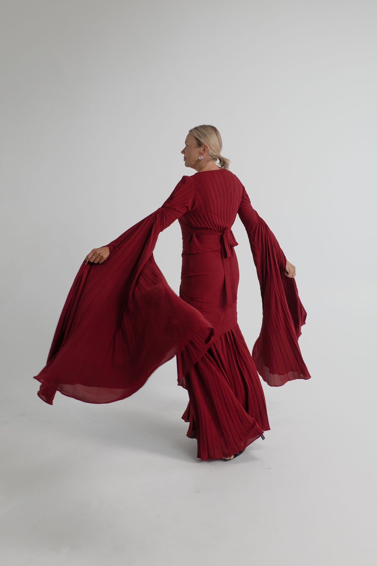Back view of the Georgia Hardinge Waterfall merlot gown in motion, showing the waist tie and flowing pleated sleeves.
