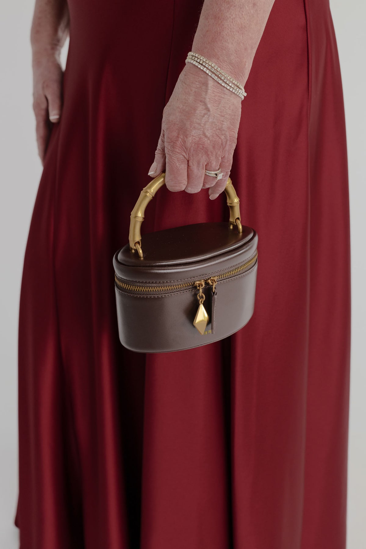 Close-up of the JW Pei Natalie dark brown top handle bag with gold-tone bamboo-style handle, zip closure and geometric pendant, styled against the Anna Quan Gabrielle wine gown.
