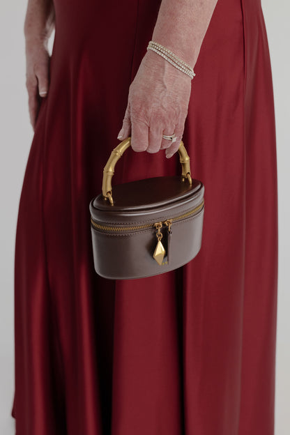 Close-up of the JW Pei Natalie dark brown top handle bag with gold-tone bamboo-style handle, zip closure and geometric pendant, styled against the Anna Quan Gabrielle wine gown.
