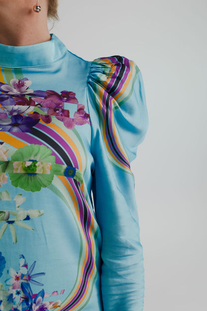 Close-up of the Mackenzie Mode Hollywood Blooming Mini Dress showing the turquoise satin finish, high neckline, gathered puff shoulder and vivid floral-and-multicolour stripe print. 
