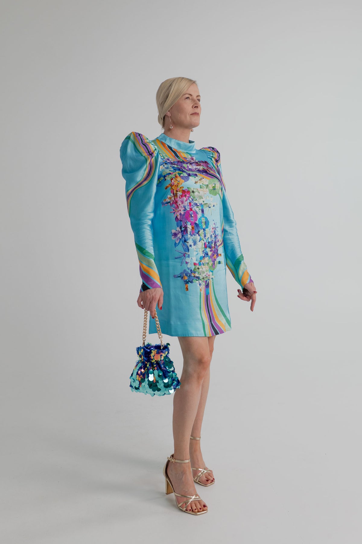 Mackenzie Mode Hollywood Blooming Mini Dress in turquoise satin with a high neckline, long puff sleeves and multicolour floral-and-stripe print, styled with the Olga Berg Pedro Sequin Pouch. 
