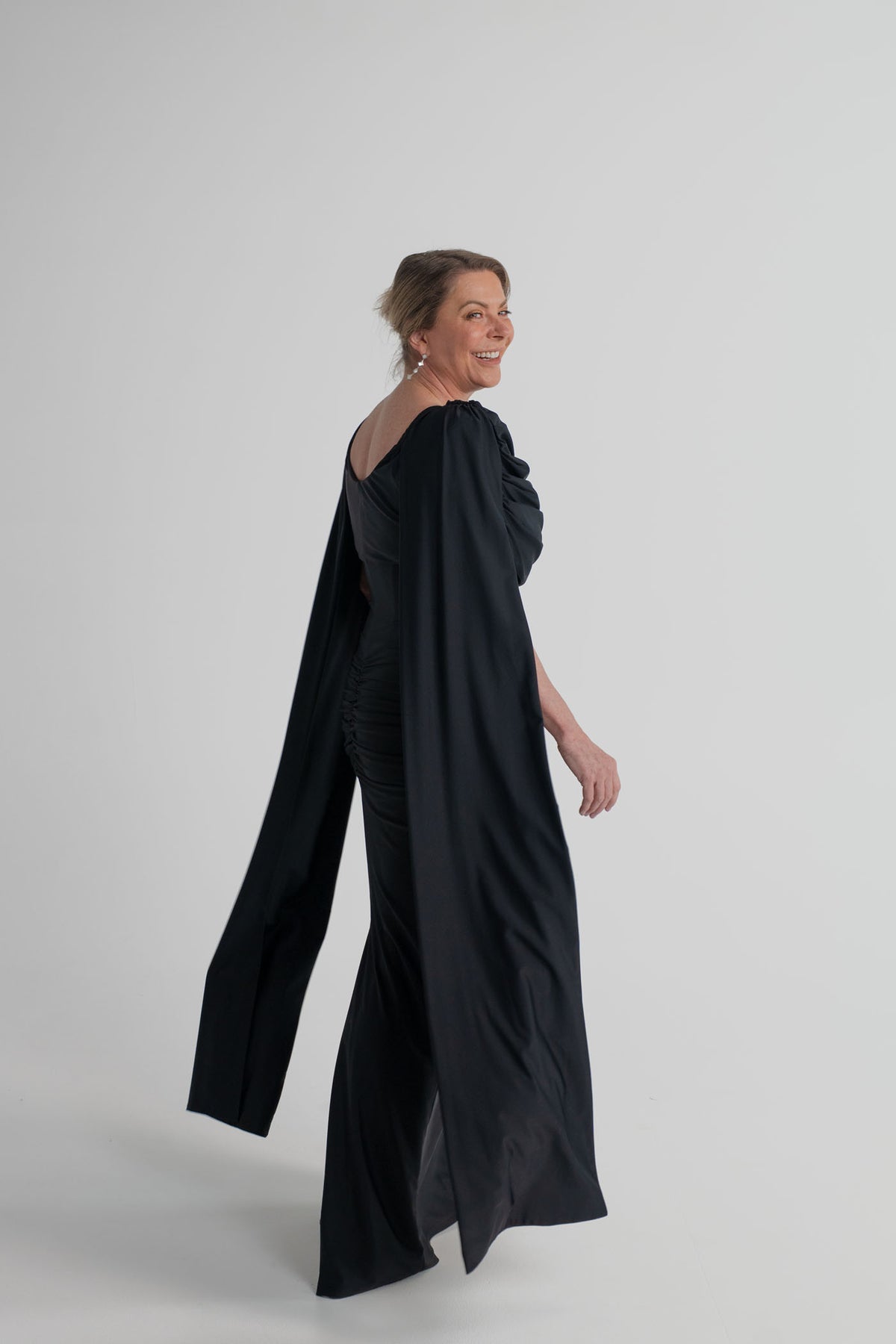 Side and back view of the Mossman Reign black midi dress showing the elongated cape sleeves, open back and fitted silhouette.
