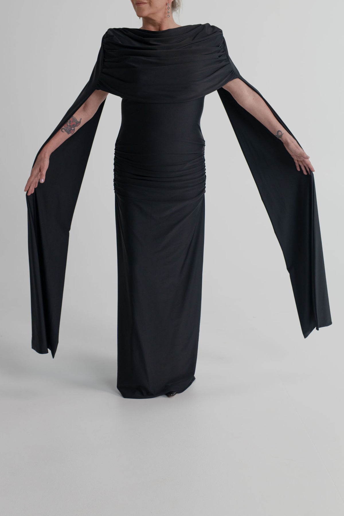 Detail of the Mossman Reign black dress showing the softly draped neckline, ruched waist and dramatic elongated cape sleeves.
