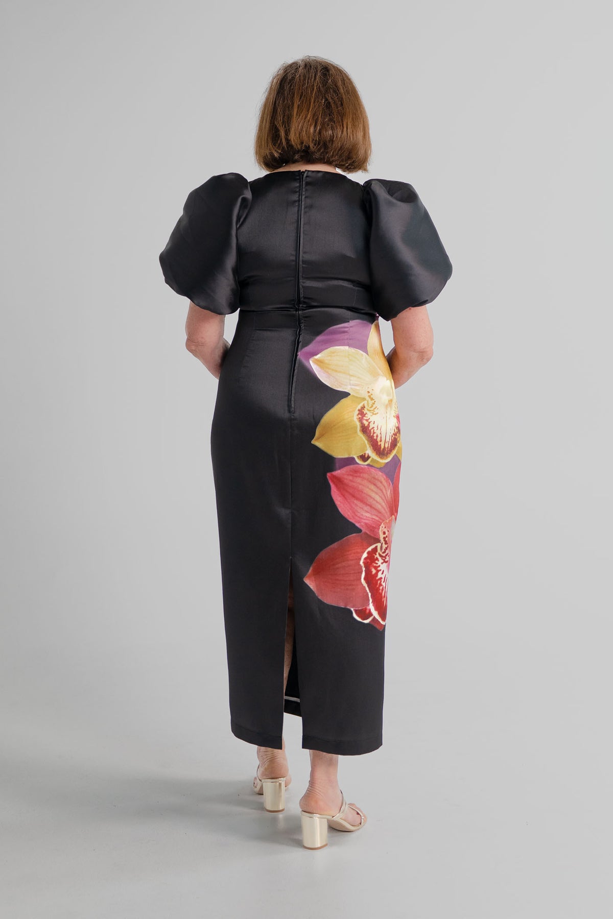 Back view of the Pasduchas Nova Black Orchid midi dress showing the puff sleeves, fitted silhouette, centre-back zip and back split.
