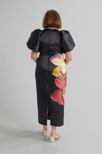 Back view of the Pasduchas Nova Black Orchid midi dress showing the puff sleeves, fitted silhouette, centre-back zip and back split.
