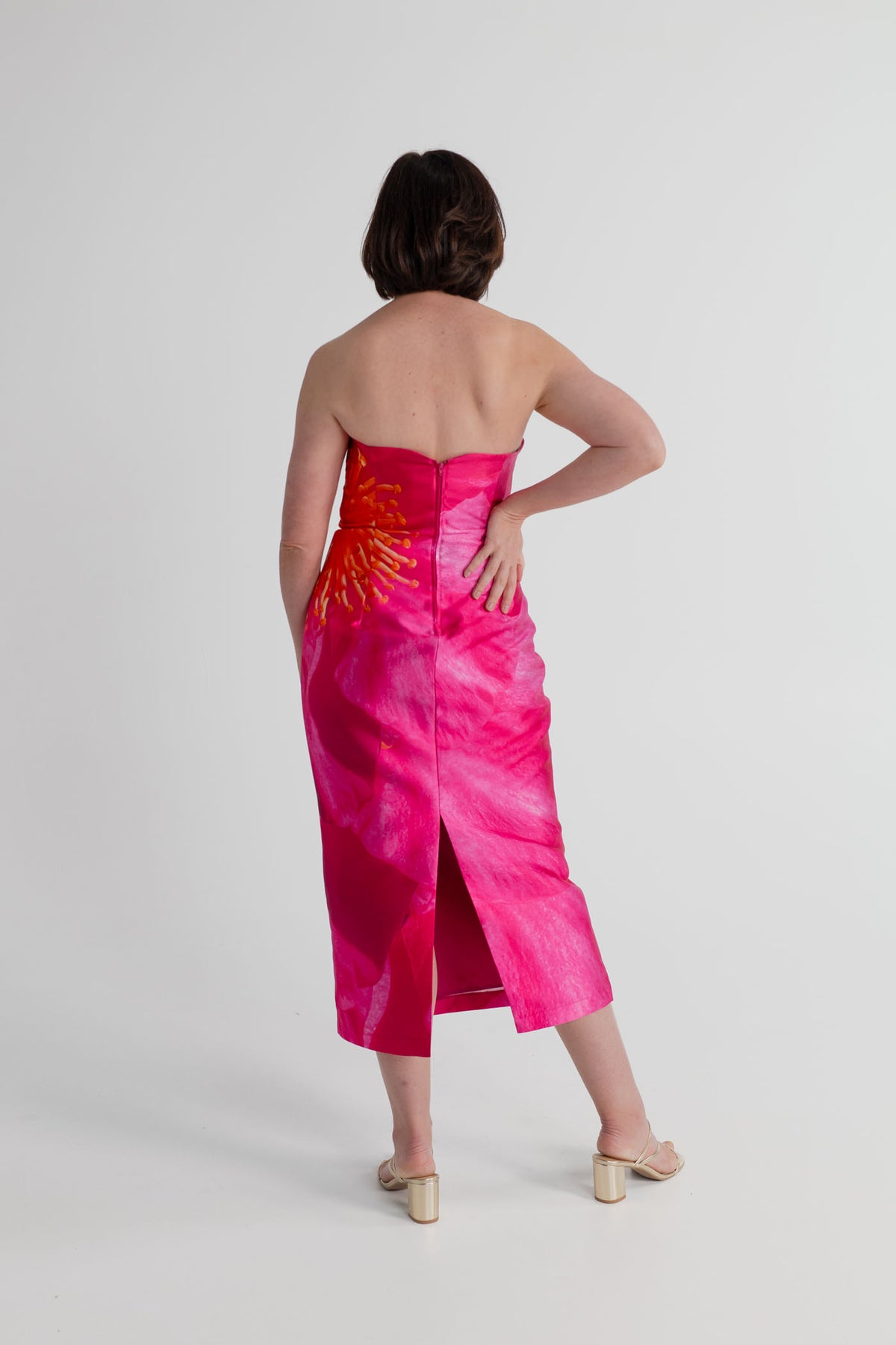 Back view of the Pasduchas Thea Camellia Pink strapless midi dress showing the fitted bodice, centre-back zip and back split.
