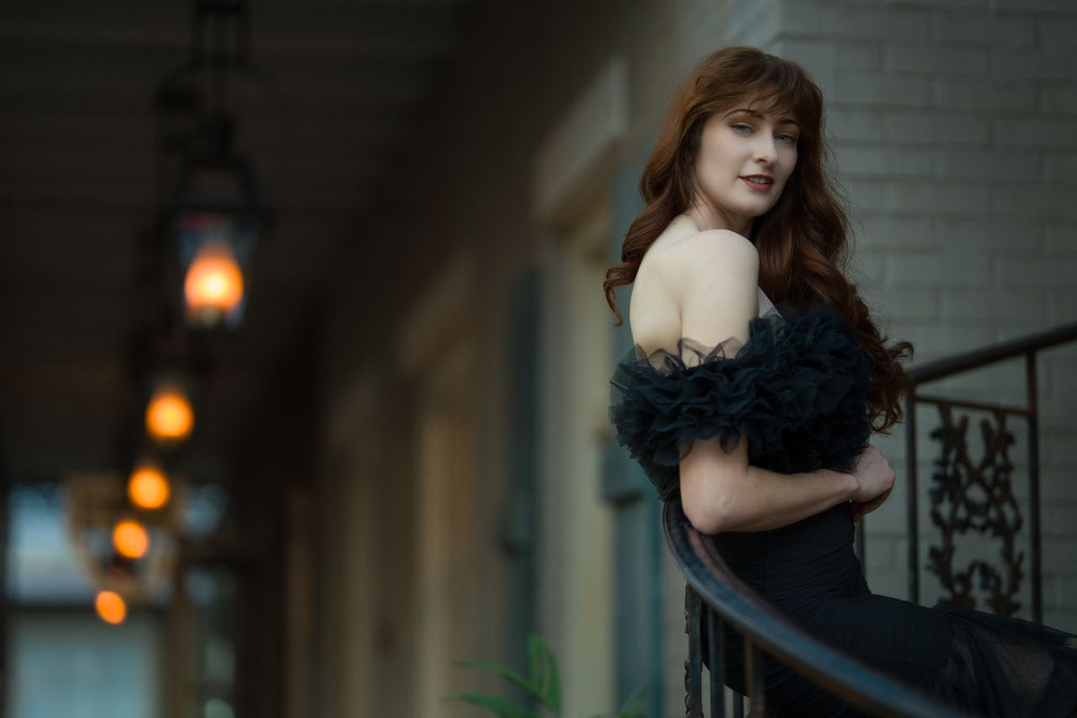 A woman with long red hair wearing a black off-the-shoulder ruffled dress, leaning against a balcony railing in a softly lit outdoor corridor.