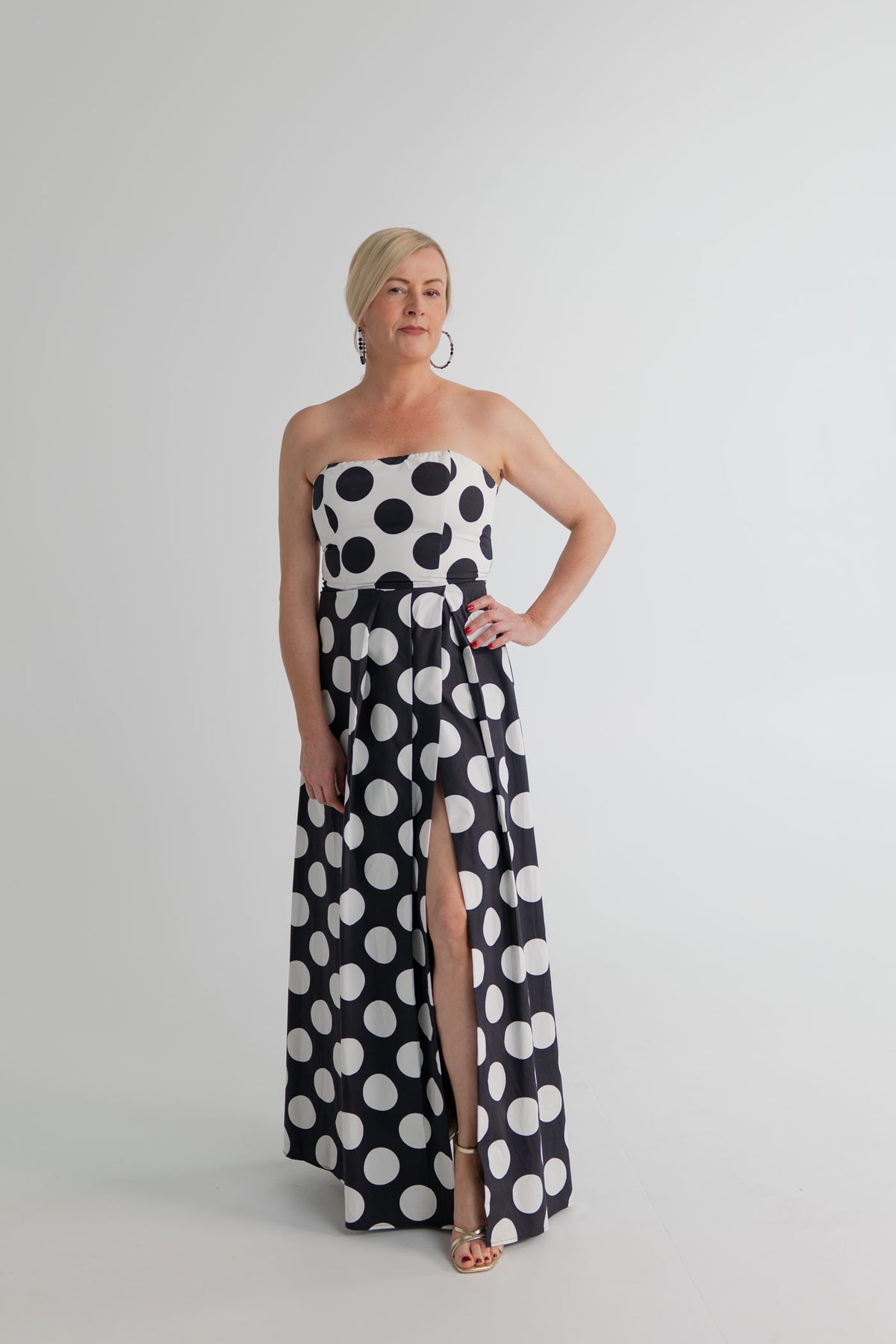 Front view of the Sonya Moda Milano Gown showing the black and white polka dot design, structured strapless bodice and high front split in the full-length skirt. 
