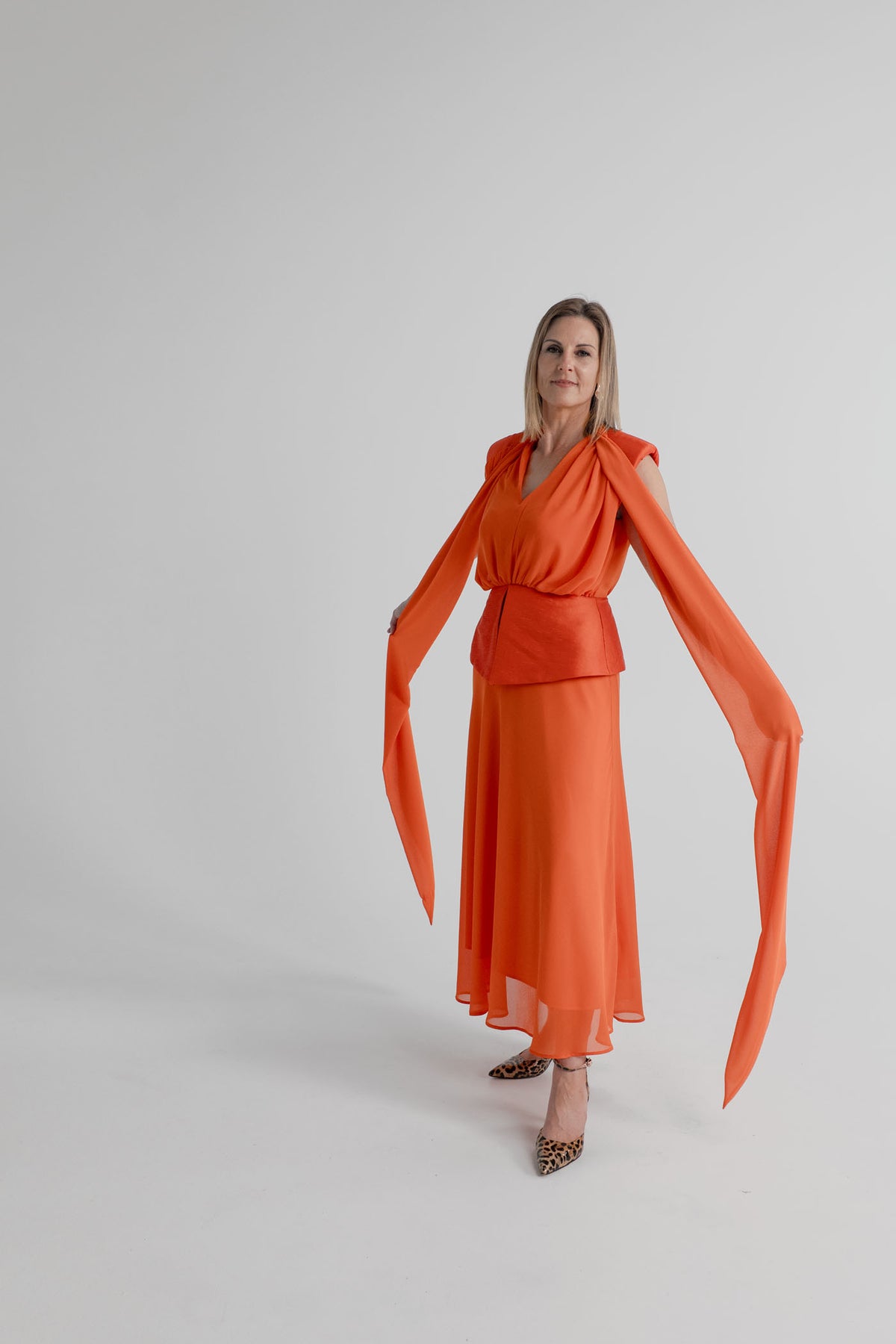 Angled front view of the orange Strena Diana Midi Dress showing the flowing shoulder scarf ties, V neckline and fitted waist detail.
