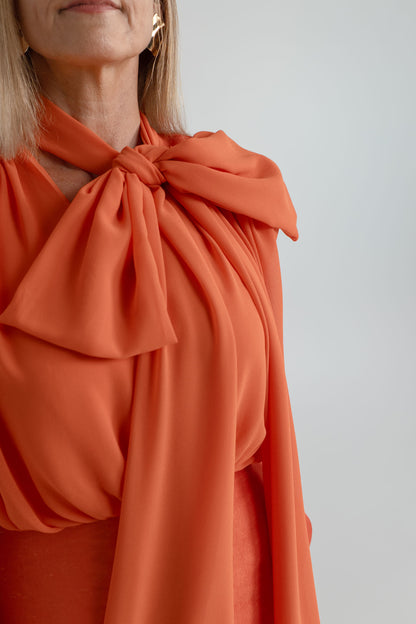 Close-up of the Strena Diana Midi Dress showing the soft orange fabric, gathered shoulders and oversized scarf tie at the neckline.
