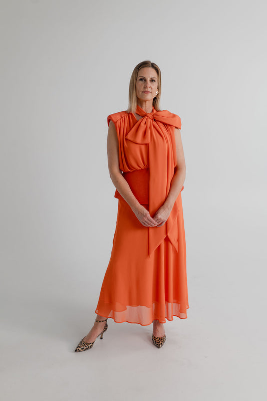 Front view of the Strena Diana Midi Dress in orange styled with the scarf tied at the neck in a statement bow.
