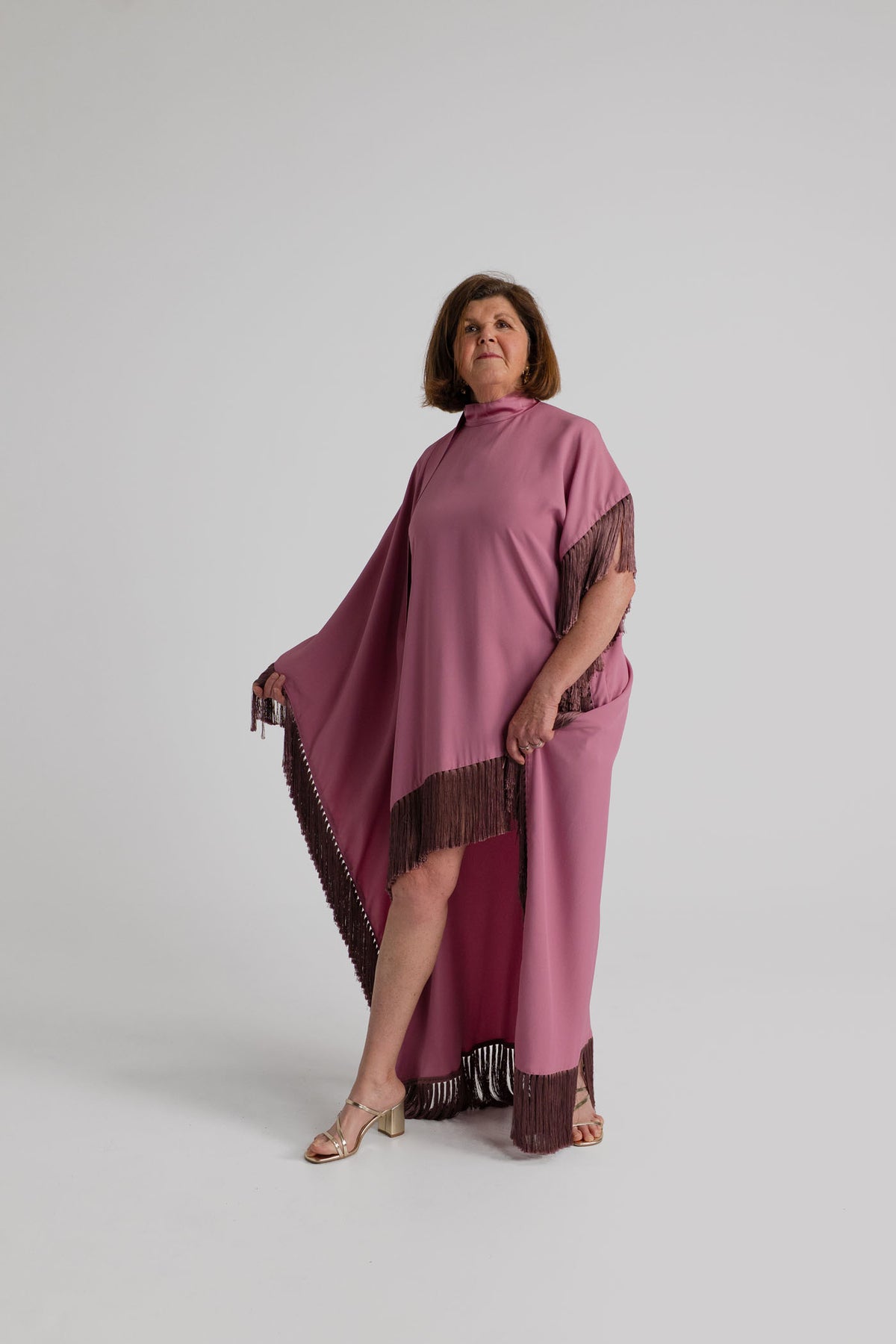 Taller Marmo Asymmetrical Fringed Satin Kaftan in dusty pink with high neckline, dramatic asymmetrical cape silhouette and dark contrast fringe, shown full length from the front.
