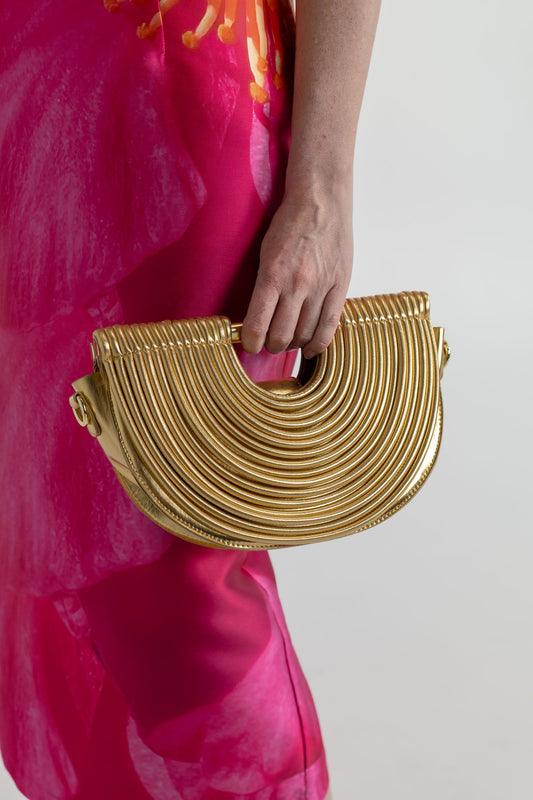Close-up of the Verano Hill Amara gold handbag with sculptural ribbed semicircle design, styled against the Pasduchas Thea Camellia Pink dress.
